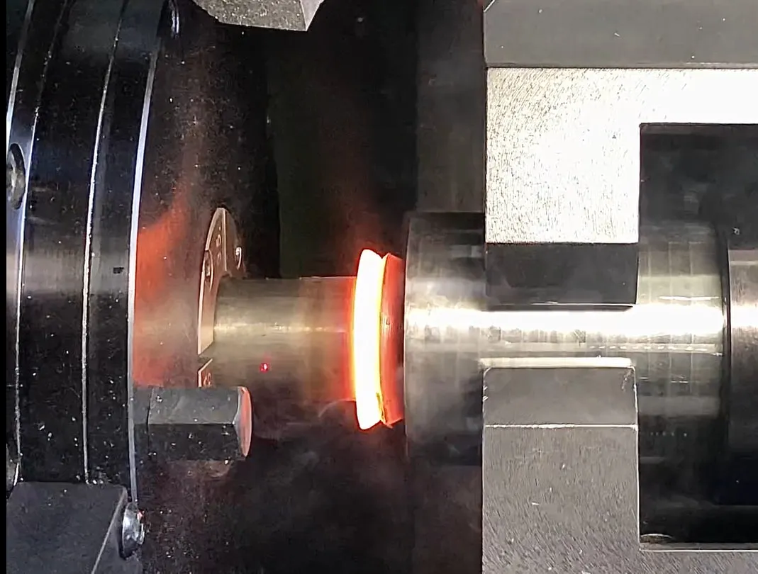 Glowing, orange-red weld on the clamped workpiece: the chuck of the rotating side on the left, the vice of the stationary side on the right